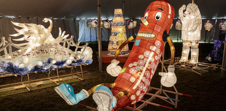 Colorful, illuminated lanterns under a tent, including a large, smiling sausage-shaped character, a swan, an elephant, and other decorative designs on display at a festival or exhibition.