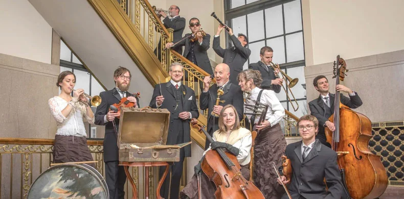 A group of musicians in formal attire poses on a staircase, playing various instruments including violin, trumpet, cello, drums, upright bass, and clarinet, with some standing and some seated, in a bright, elegant indoor setting.