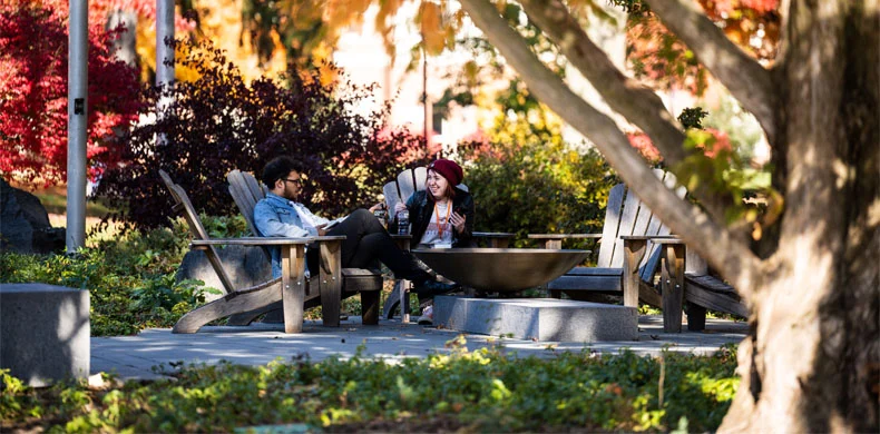 Two people sit on wooden chairs in a leafy outdoor area, engaged in conversation. Autumn trees with colorful leaves surround them, creating a peaceful atmosphere.
