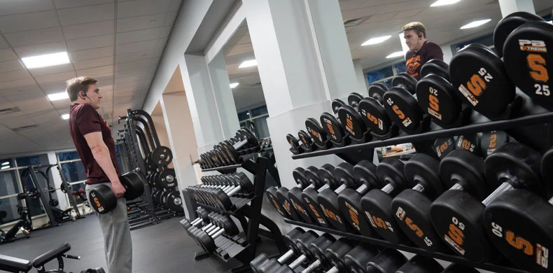 A person in a gym holds a dumbbell near a rack of weights, looking at their reflection in a large wall mirror. The gym has bright lighting and various exercise equipment.