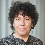 A woman with short, curly dark hair and light skin is wearing dangly earrings and a patterned top. She is smiling softly and standing in front of a blurred, light-colored background.