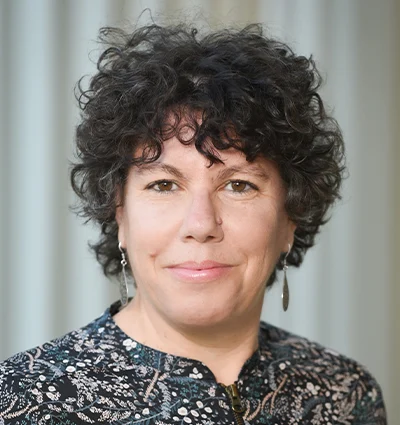 A woman with short, curly dark hair and light skin is wearing dangly earrings and a patterned top. She is smiling softly and standing in front of a blurred, light-colored background.
