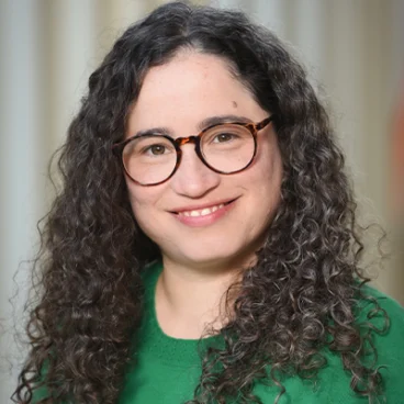 A woman with long, curly dark hair and glasses is smiling. She is wearing a green sweater and standing in front of a softly blurred, light-colored background.