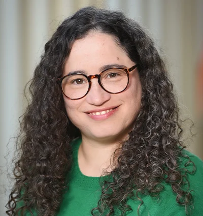 A woman with long, curly dark hair and glasses is smiling. She is wearing a green sweater and standing in front of a softly blurred, light-colored background.