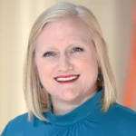 A smiling woman with straight blonde hair wearing a teal high-neck top stands in front of a softly blurred beige and orange background.