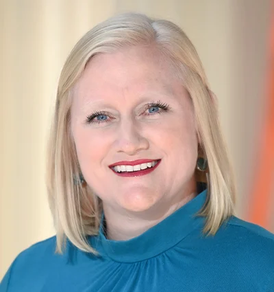 A smiling woman with straight blonde hair wearing a teal high-neck top stands in front of a softly blurred beige and orange background.