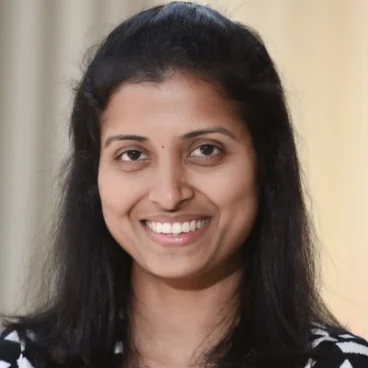 A woman with long dark hair is smiling, wearing a black and white striped top, standing in front of a neutral, blurred background.