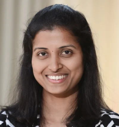 A woman with long dark hair is smiling, wearing a black and white striped top, standing in front of a neutral, blurred background.