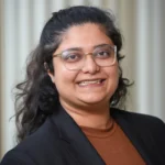 A person with long, wavy dark hair and glasses smiles at the camera. They are wearing a black blazer over a brown top, standing in front of a softly blurred, light-colored background with vertical lines.