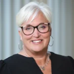 Smiling woman with short white hair, wearing glasses, silver hoop earrings, and a black top, standing in front of a softly blurred light-colored background.