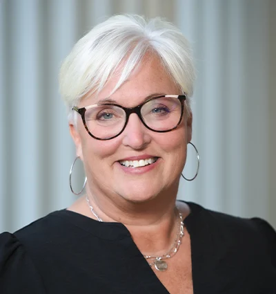 Smiling woman with short white hair, wearing glasses, silver hoop earrings, and a black top, standing in front of a softly blurred light-colored background.