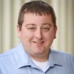 A man with short brown hair and light skin smiles at the camera. He is wearing a light blue collared shirt over a white undershirt. The background is softly blurred with vertical beige stripes.