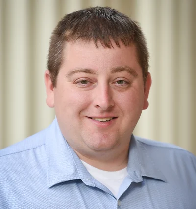 A man with short brown hair and light skin smiles at the camera. He is wearing a light blue collared shirt over a white undershirt. The background is softly blurred with vertical beige stripes.