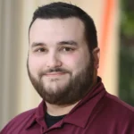 A man with short dark hair and a beard, wearing a burgundy collared shirt, smiles softly while standing in front of a blurred background with neutral tones and some greenery.