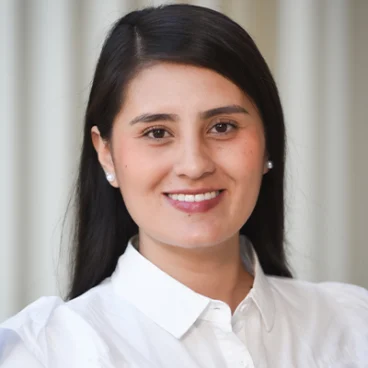 A woman with long dark hair, wearing a white button-up shirt and pearl earrings, smiles at the camera. The background features soft, blurred vertical lines in neutral tones.
