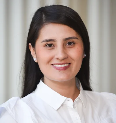 A woman with long dark hair, wearing a white button-up shirt and pearl earrings, smiles at the camera. The background features soft, blurred vertical lines in neutral tones.