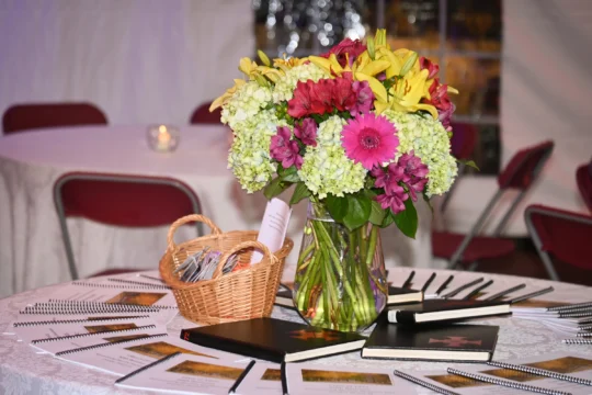 A round table with a colorful bouquet of flowers in a glass vase, a small basket filled with pens, and several notebooks and spiral-bound booklets arranged in a fan shape—perfect for a Homecoming-Reunion gathering. Red chairs and a candle are in the background.