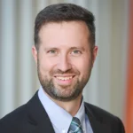 A man with short brown hair and a beard, wearing a dark suit, light blue shirt, and patterned tie, smiles at the camera in front of a blurred background with vertical lines.