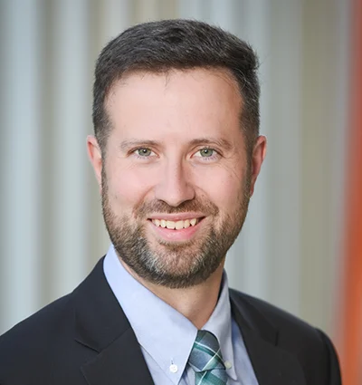 A man with short brown hair and a beard, wearing a dark suit, light blue shirt, and patterned tie, smiles at the camera in front of a blurred background with vertical lines.