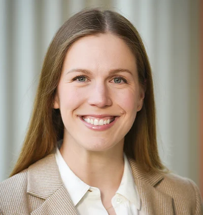 A woman with straight, light brown hair and a friendly smile is wearing a tan blazer over a white collared shirt. The background is softly blurred with vertical lines.