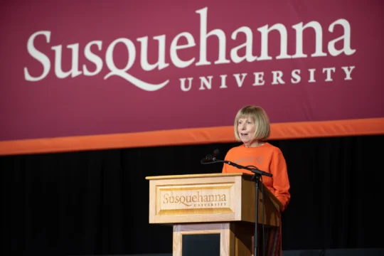 A woman wearing an orange top speaks at a podium with the 体育买球 University logo, in front of a large maroon 体育买球 University banner during a Homecoming-Reunion event.