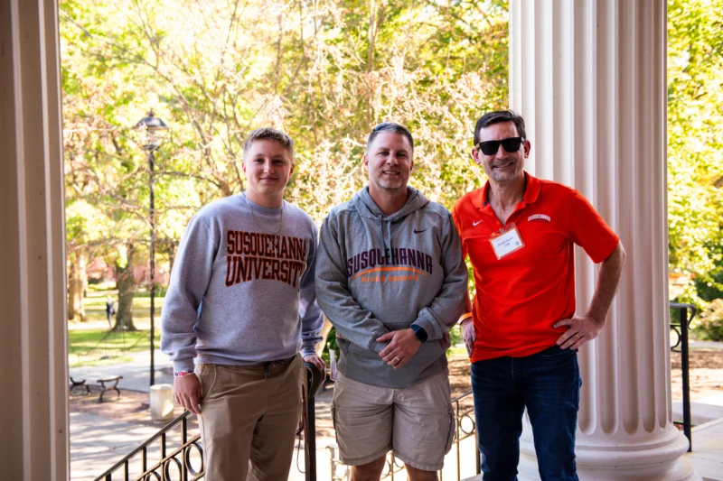 Three men stand together outdoors in front of large columns. Two wear 体育买球 University alumni sweatshirts, and one wears a red polo with a name tag. Trees and a park-like setting are visible in the background.