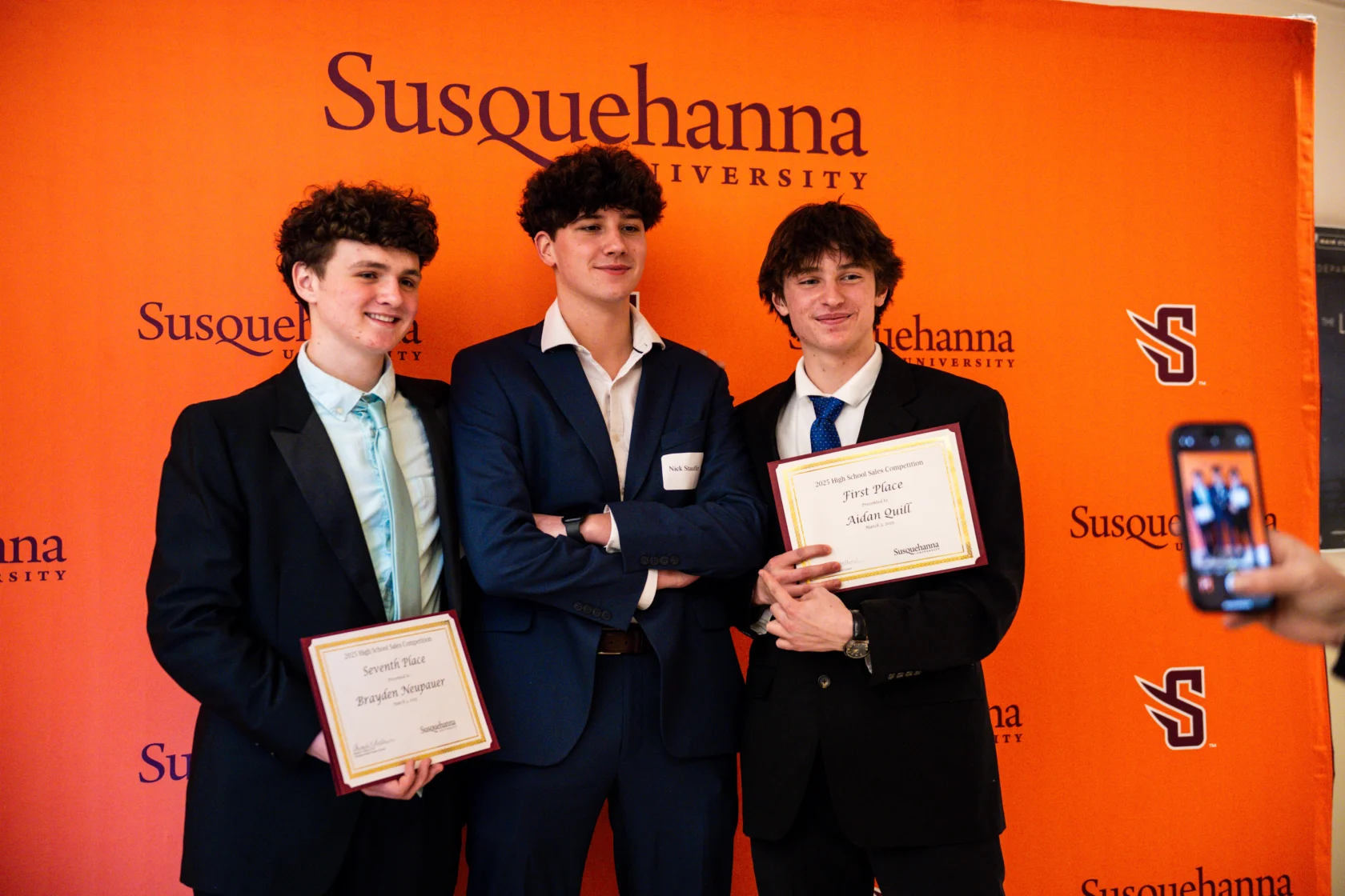 Three young men in suits stand in front of an orange 体育买球 University backdrop; two hold certificates while the third stands with arms crossed. Someone in the foreground snaps a photo with a phone.