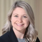 A woman with long, wavy blonde hair smiles at the camera. She is wearing a black blazer over a gray top, and the background is softly blurred in neutral tones.