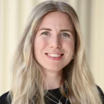 A woman with long, wavy blonde hair smiles at the camera. She is wearing a black top and layered silver necklaces. The background is softly blurred in beige tones.