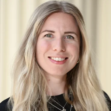 A woman with long, wavy blonde hair smiles at the camera. She is wearing a black top and layered silver necklaces. The background is softly blurred in beige tones.