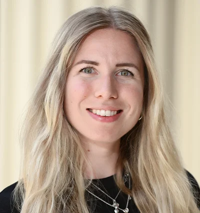 A woman with long, wavy blonde hair smiles at the camera. She is wearing a black top and layered silver necklaces. The background is softly blurred in beige tones.