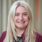 A woman with long, straight blonde hair smiles at the camera. She is wearing a maroon jacket over a patterned top, and the background is softly blurred with vertical lines.
