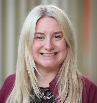 A woman with long, straight blonde hair smiles at the camera. She is wearing a maroon jacket over a patterned top, and the background is softly blurred with vertical lines.