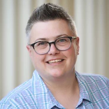 A person with short, spiked hair and glasses smiles at the camera. They are wearing a blue and purple plaid shirt. The background is softly blurred with vertical beige lines.
