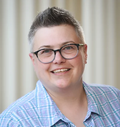 A person with short, spiked hair and glasses smiles at the camera. They are wearing a blue and purple plaid shirt. The background is softly blurred with vertical beige lines.