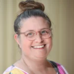A smiling woman with light skin, wearing pink-rimmed glasses and a colorful top, has her brown hair tied up in a high bun. The background is softly blurred in neutral tones.