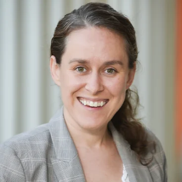 A woman with long brown hair pulled back, wearing a gray plaid blazer and white top, smiles at the camera. The background features vertical, blurred light-colored panels.