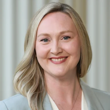 A woman with long blonde hair, wearing a light gray blazer and a cream top, smiles at the camera. The background is softly blurred with vertical light-colored lines.
