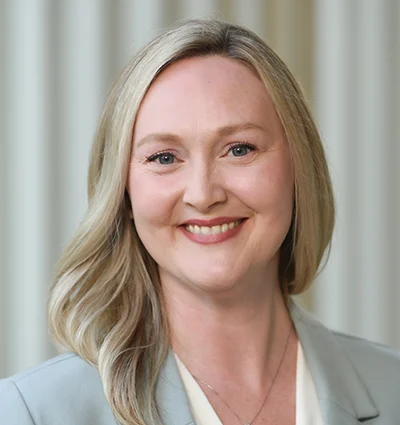 A woman with long blonde hair, wearing a light gray blazer and a cream top, smiles at the camera. The background is softly blurred with vertical light-colored lines.