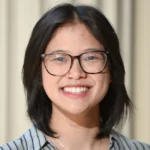 A young person with short black hair and glasses smiles at the camera. They are wearing a striped shirt and a necklace, and the background is softly blurred beige curtains.