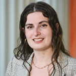 A woman with long wavy brown hair, wearing a plaid blazer, gold hoop earrings, and a delicate necklace, smiles at the camera. The background is blurred with vertical light and orange stripes.