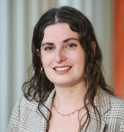 A woman with long wavy brown hair, wearing a plaid blazer, gold hoop earrings, and a delicate necklace, smiles at the camera. The background is blurred with vertical light and orange stripes.
