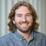 A man with wavy, shoulder-length light brown hair and a beard smiles at the camera. He is wearing a blue button-up shirt. The background is softly blurred with vertical light patterns.