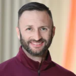 A man with short dark hair and a trimmed beard, wearing a maroon zip-up jacket, smiles at the camera with a softly blurred background.
