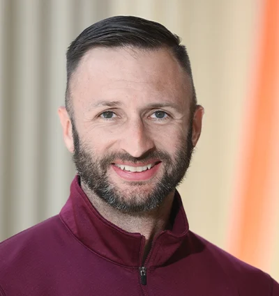 A man with short dark hair and a trimmed beard, wearing a maroon zip-up jacket, smiles at the camera with a softly blurred background.