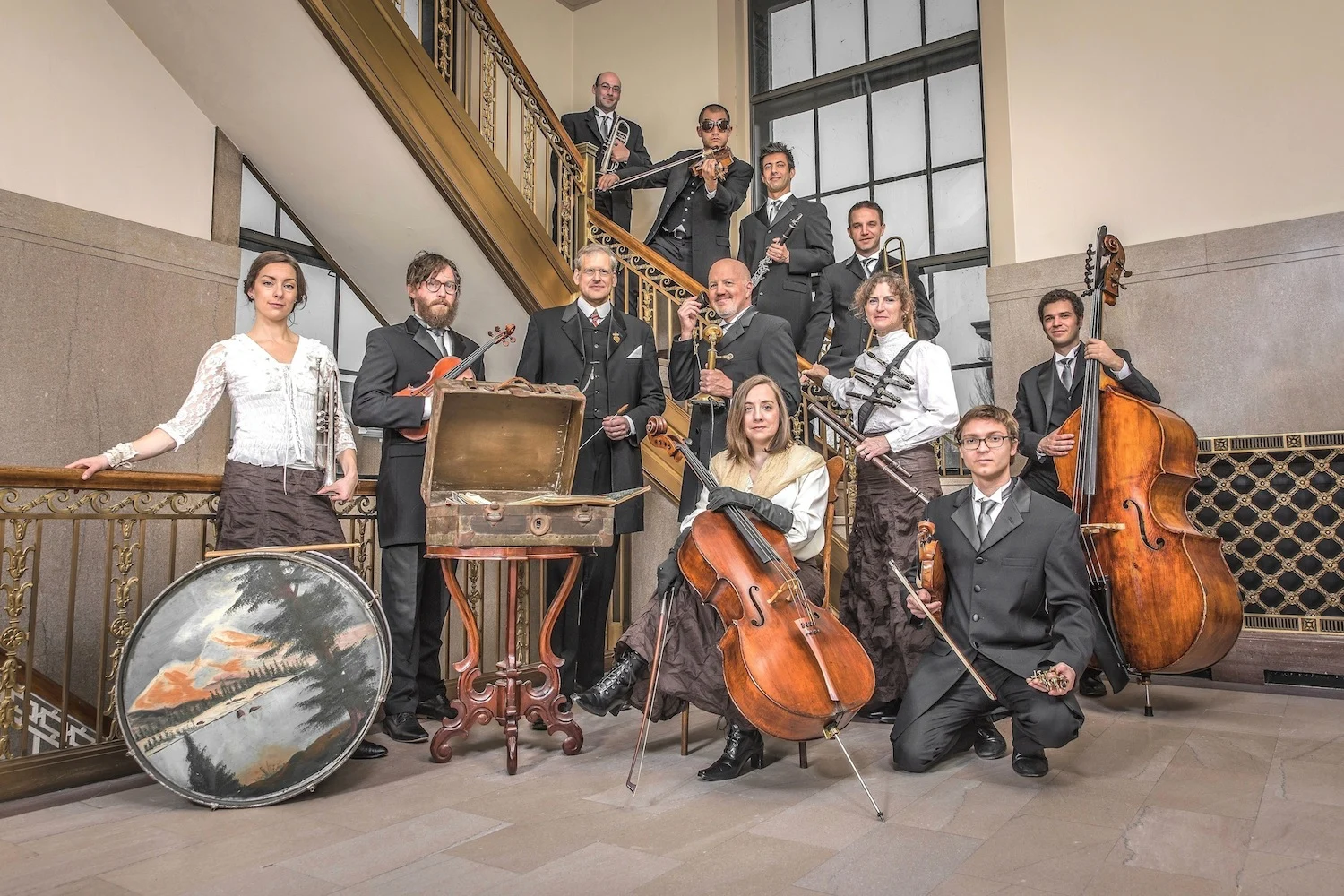 A group of eleven musicians in formal attire pose indoors with various instruments, including a drum, violin, cello, double bass, and accordion, in front of a staircase and large windows at the Symposium on Historic American Music.