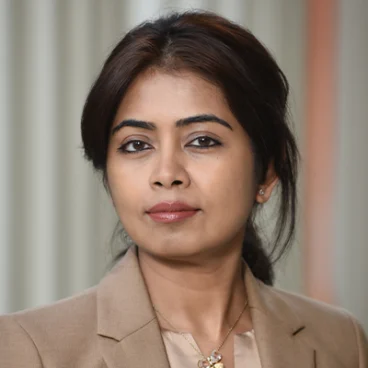 A woman with dark hair pulled back, wearing a tan blazer and a necklace with a flower pendant, looks confidently at the camera. The background is softly blurred with neutral tones.