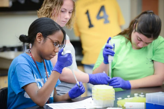 Three students in a lab use pipettes and wear purple gloves while working with yellow-tipped sample trays. Two focus on the experiment; one observes, as if in a scene at the Actor's Institute. Laboratory equipment and notes are visible on the table.