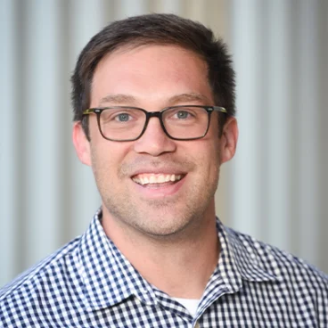 A smiling man with short brown hair and glasses wears a black and white checkered shirt, standing in front of a blurred, light-colored background.
