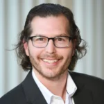 A man with wavy brown hair, glasses, and a beard smiles at the camera. He is wearing a black blazer over a white shirt, and the background is softly blurred with vertical light-colored lines.